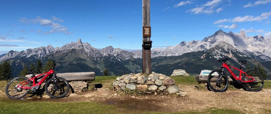 Wooden cross on mountain summit with two red bikes and Alps in background