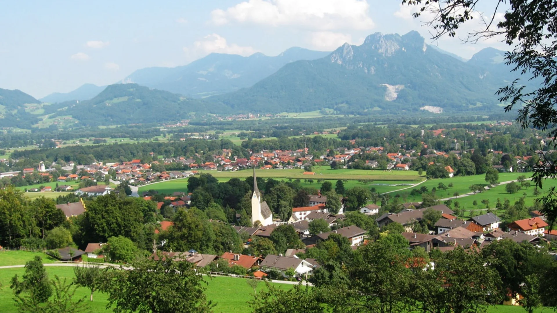 Blick auf ein Dorf mit Kirche vor grünen Wiesen und Bergen bei klarem Himmel