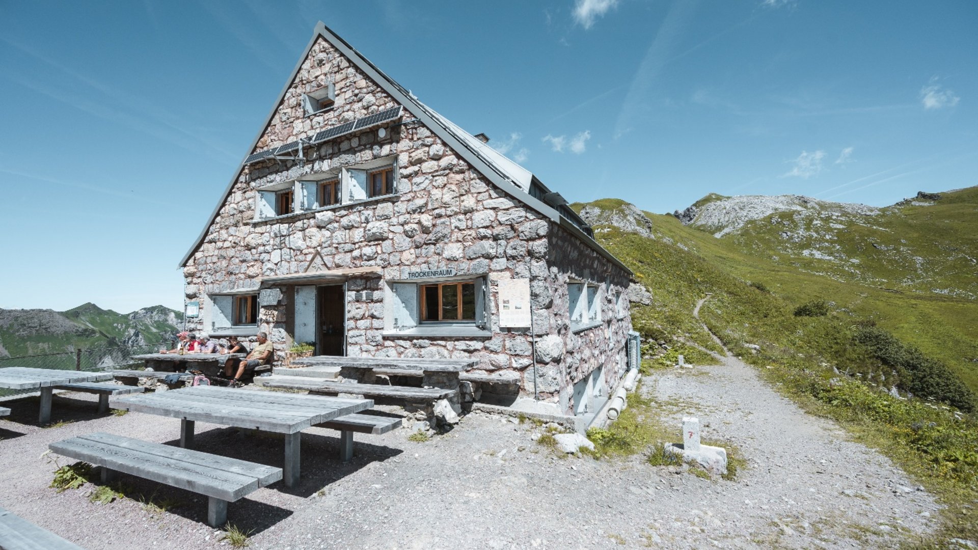 Stone house with benches and hikers in mountainous landscape under clear sky