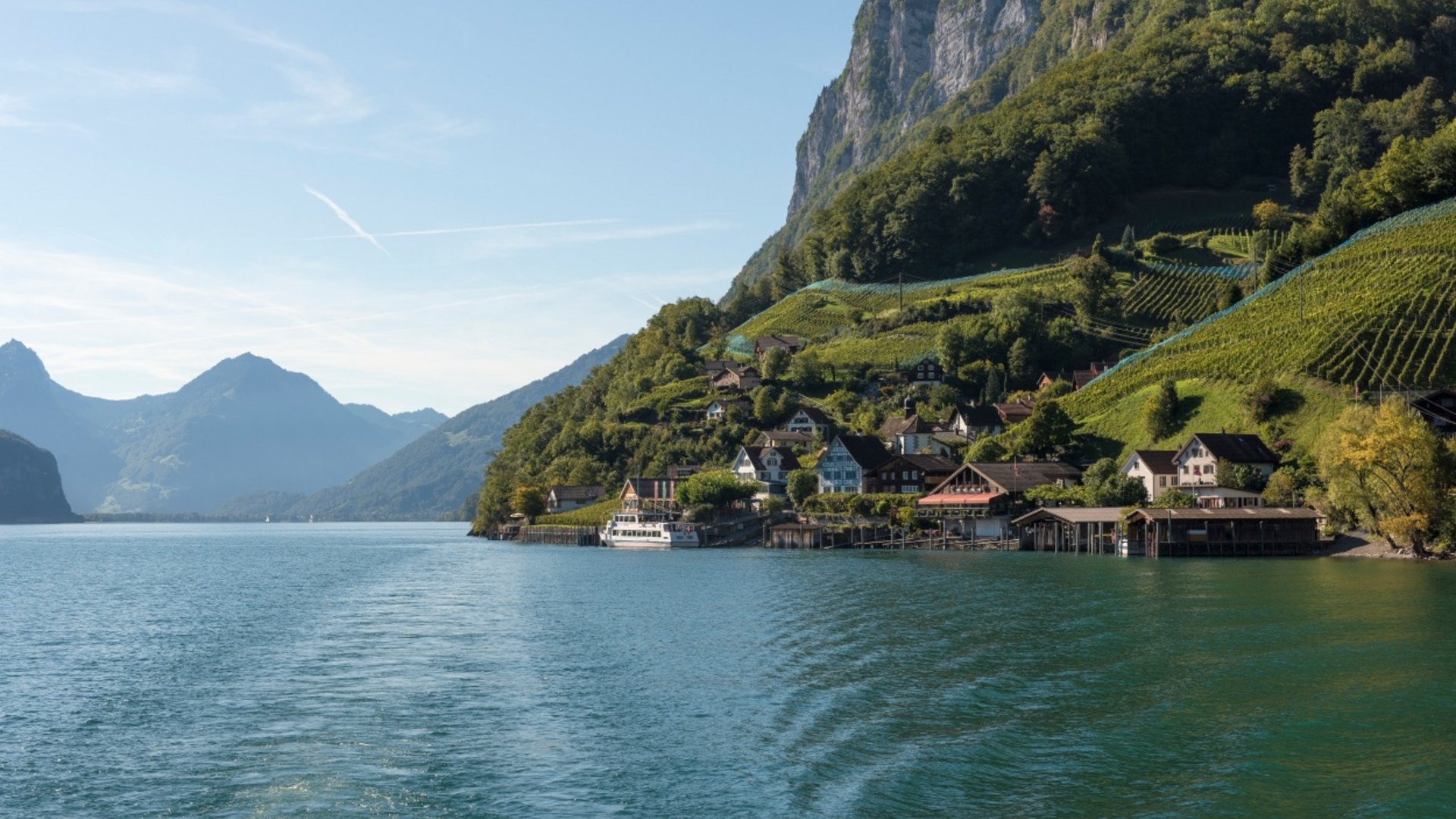 Village by lakeside beneath forested mountain on clear day