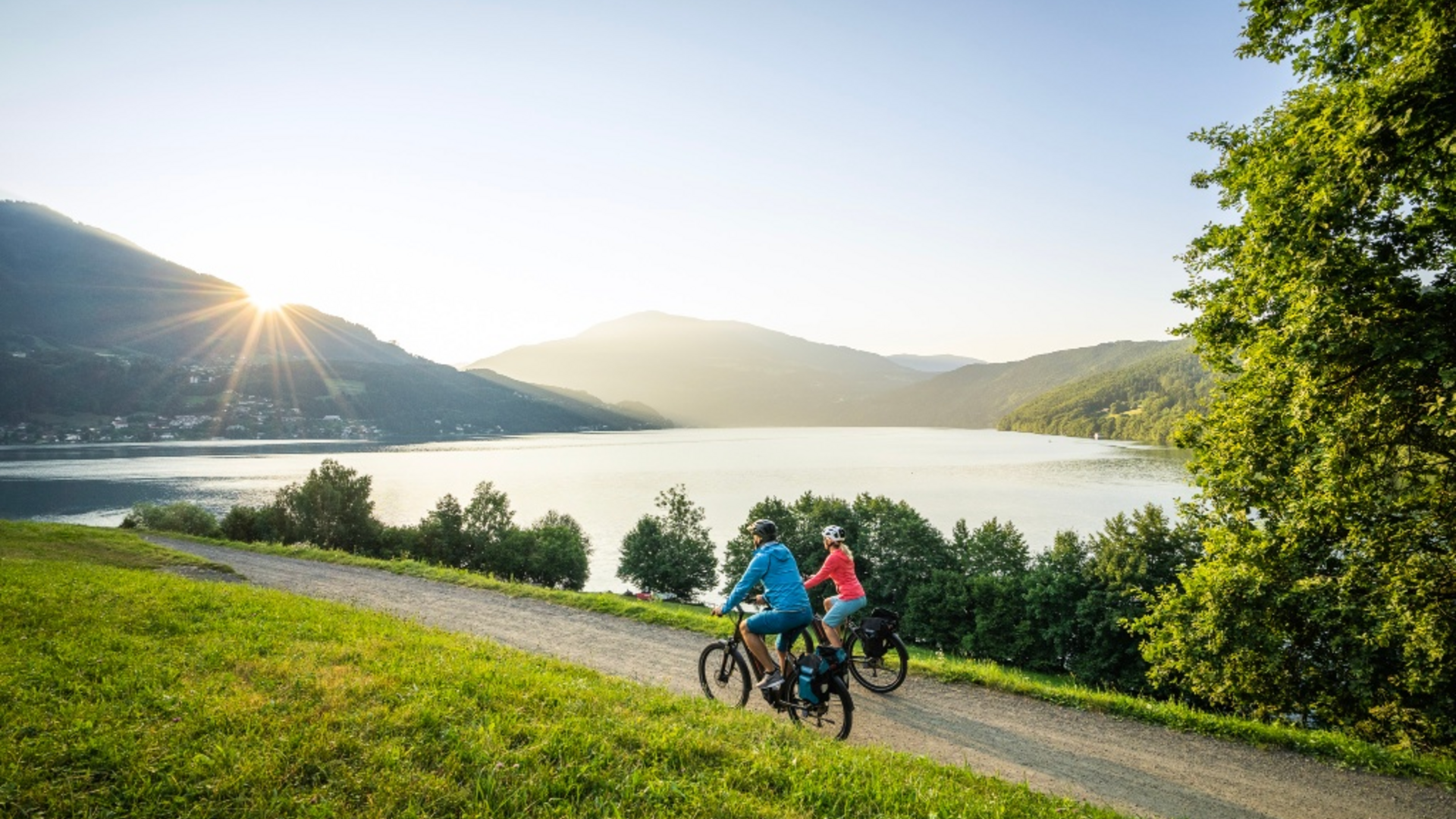 Two cyclists riding on a path by a lake with mountains and sunset