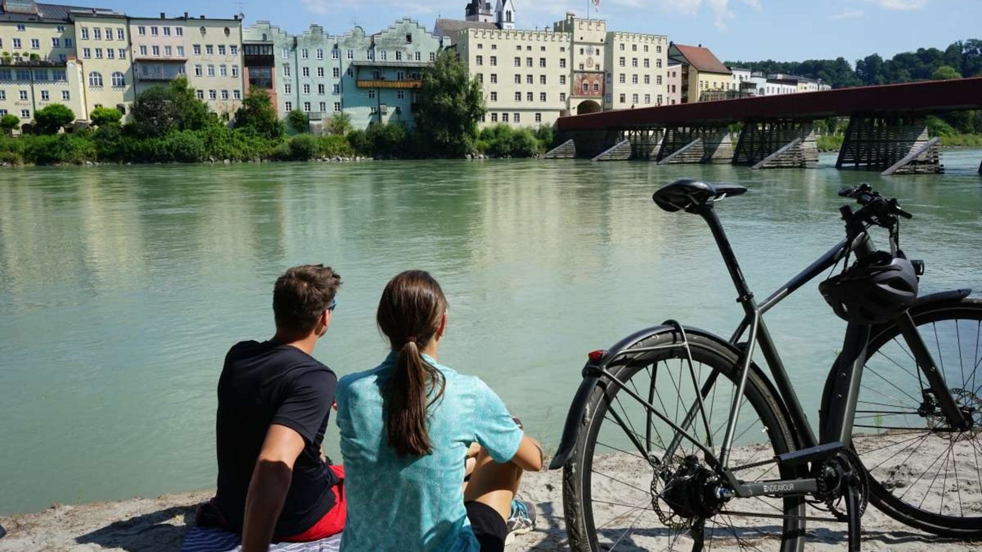 Two people sitting by the river with a bicycle and colorful buildings in view