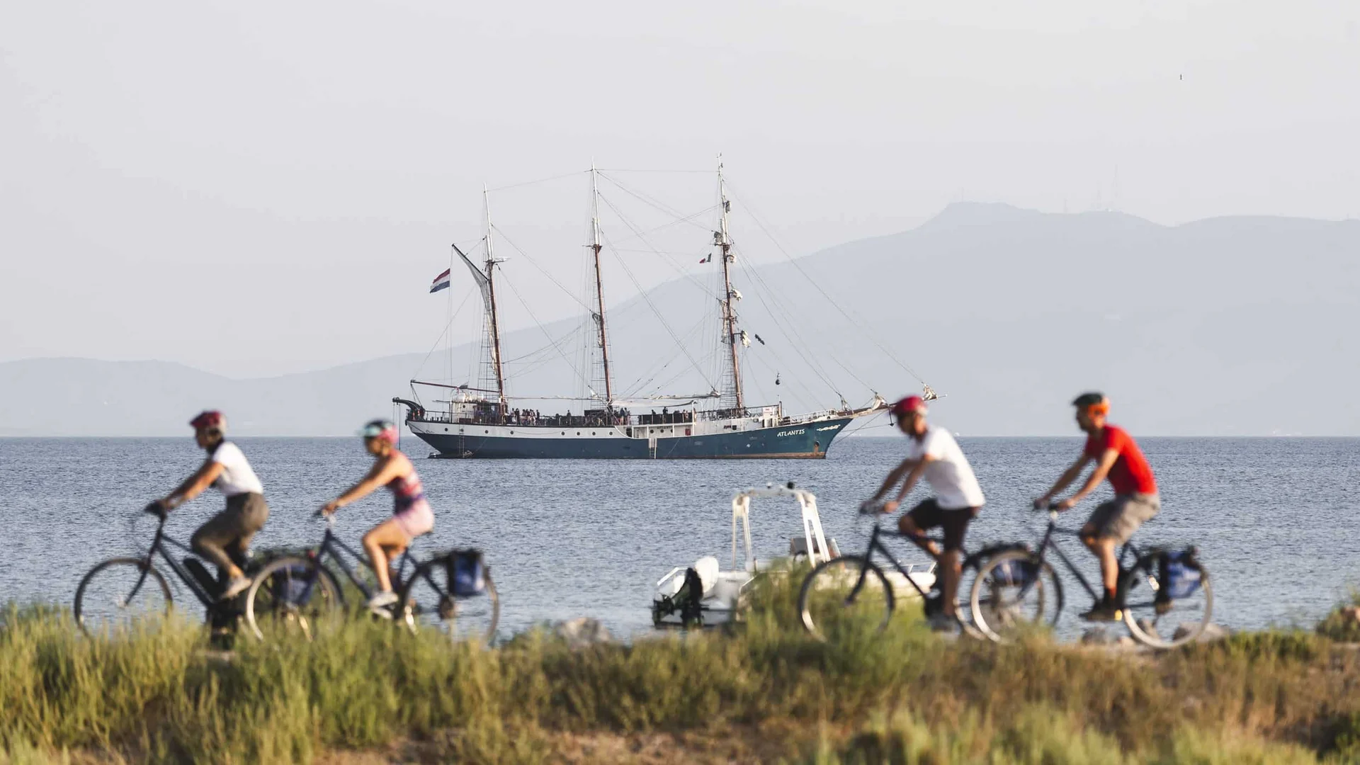 Vier Radfahrer auf Küstenweg mit Segelschiff und Bergen im Hintergrund