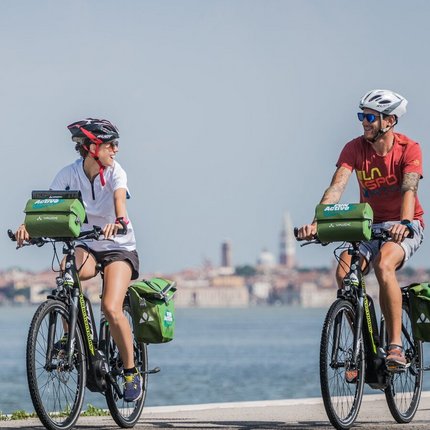 Two cyclists wearing helmets with green bags ride along a waterfront path