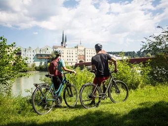 Chiemsee-Alpenland © Velontour Two cyclists wearing helmets overlooking riverside buildings on a sunny day