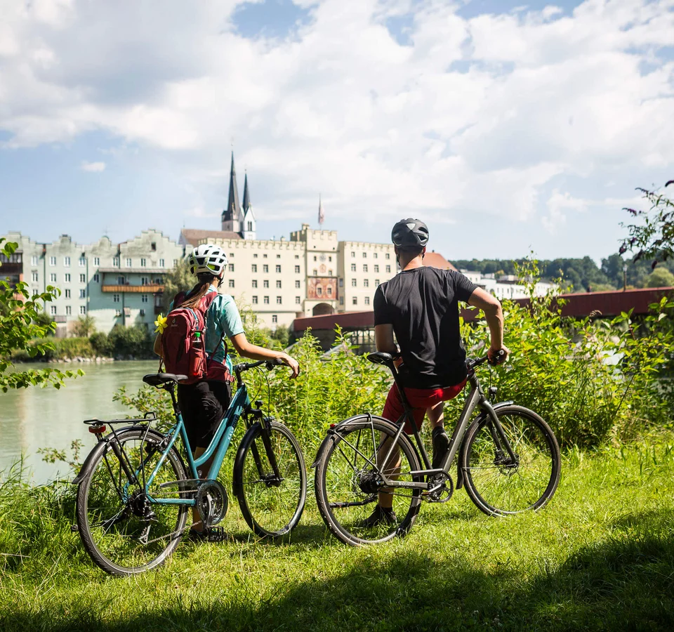 Zwei Radfahrer mit Helm blicken auf Gebäude am Flussufer an einem sonnigen Tag