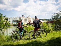 Two cyclists wearing helmets overlooking riverside buildings on a sunny day