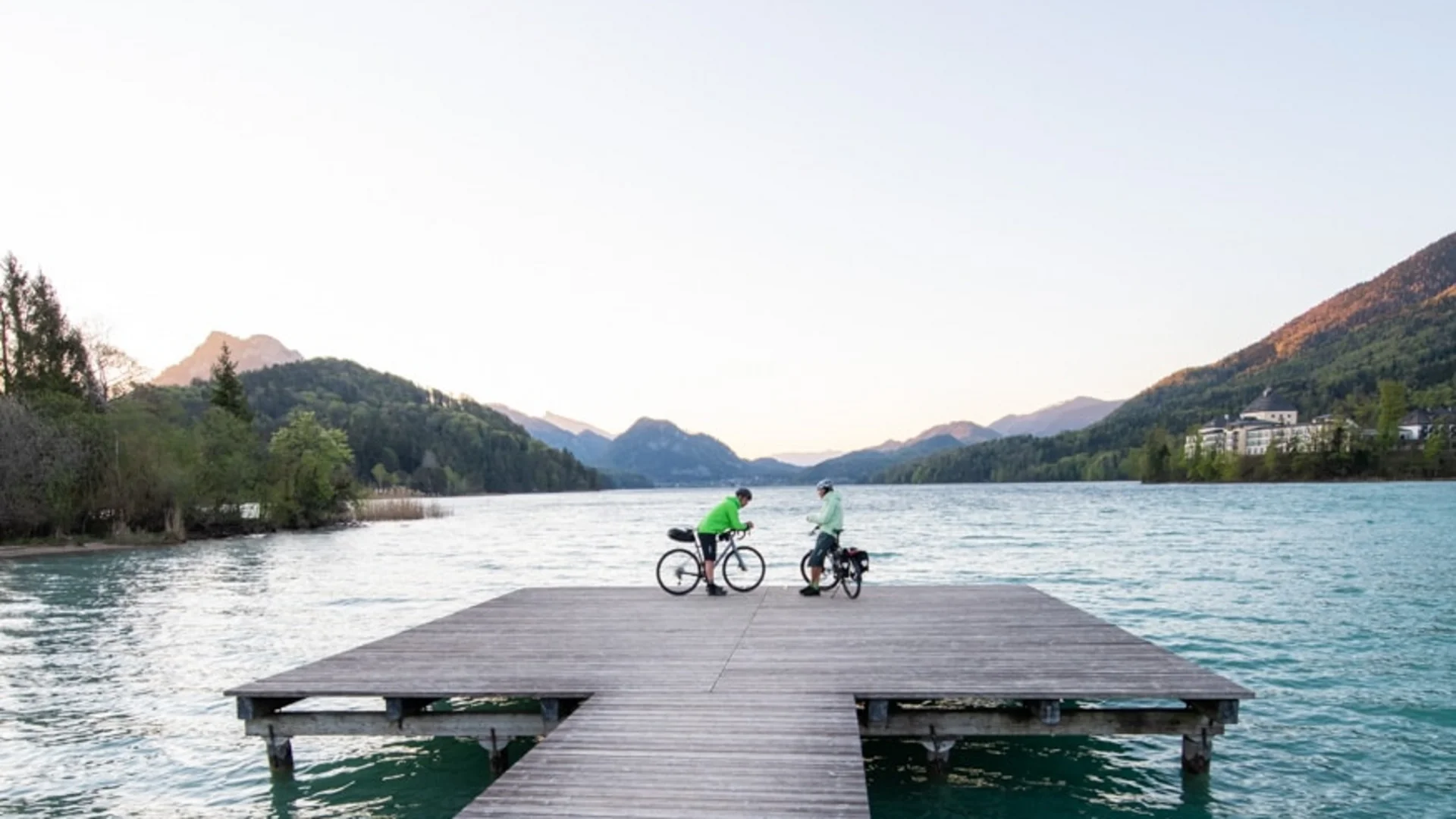 Two cyclists on a wooden pier over a lake with mountains in the background
