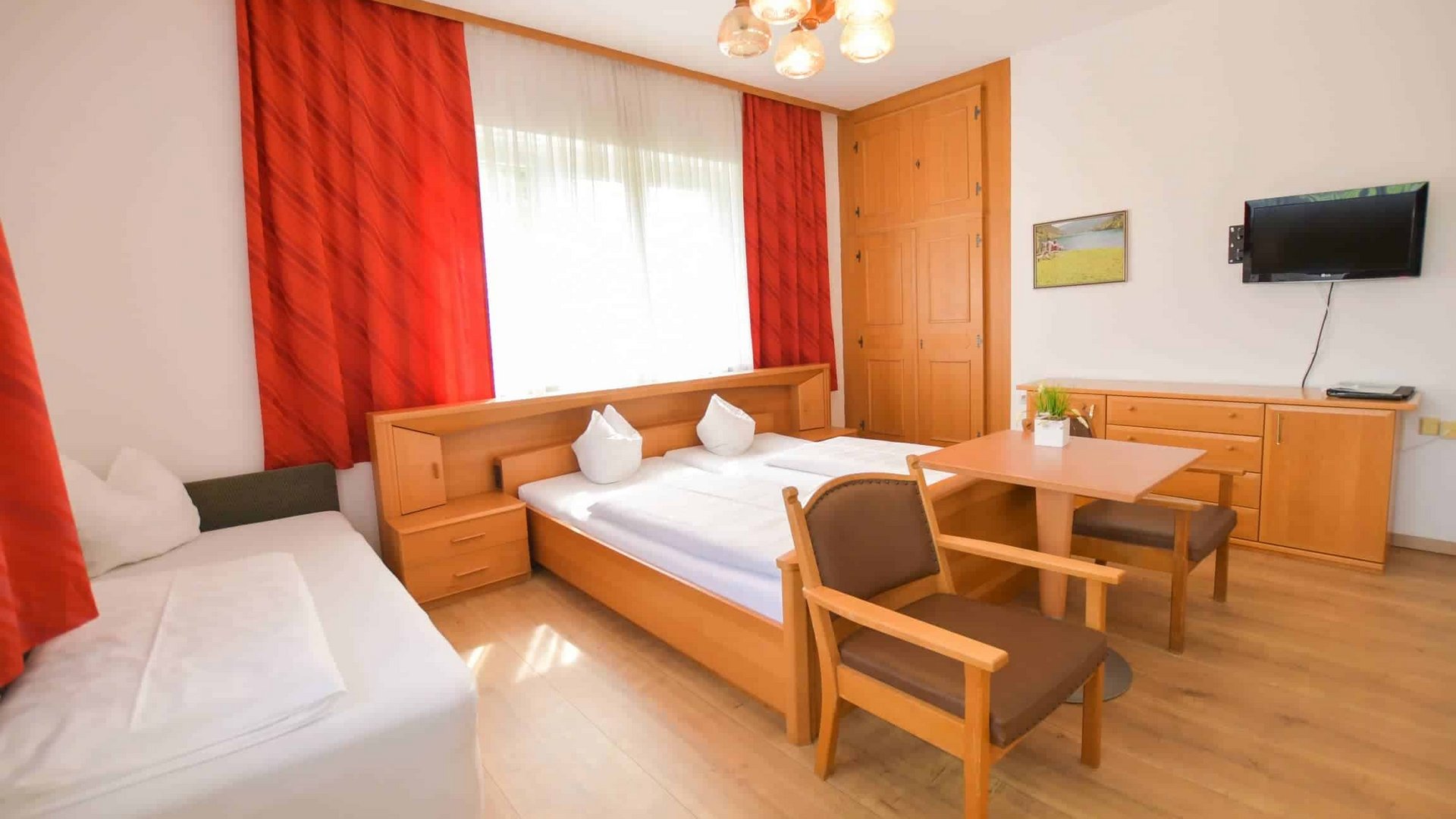 Bright hotel room with double bed, sofa, table, chairs, and red curtains