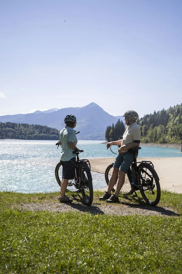 Die schönsten Radregionen im Süden Deutschlands © Velontour - Heiko Mandl Zwei Radfahrer mit Helm machen Pause an einem See mit Bergen im Hintergrund