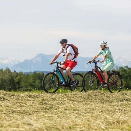 Two people cycling on a field path with mountains in the background