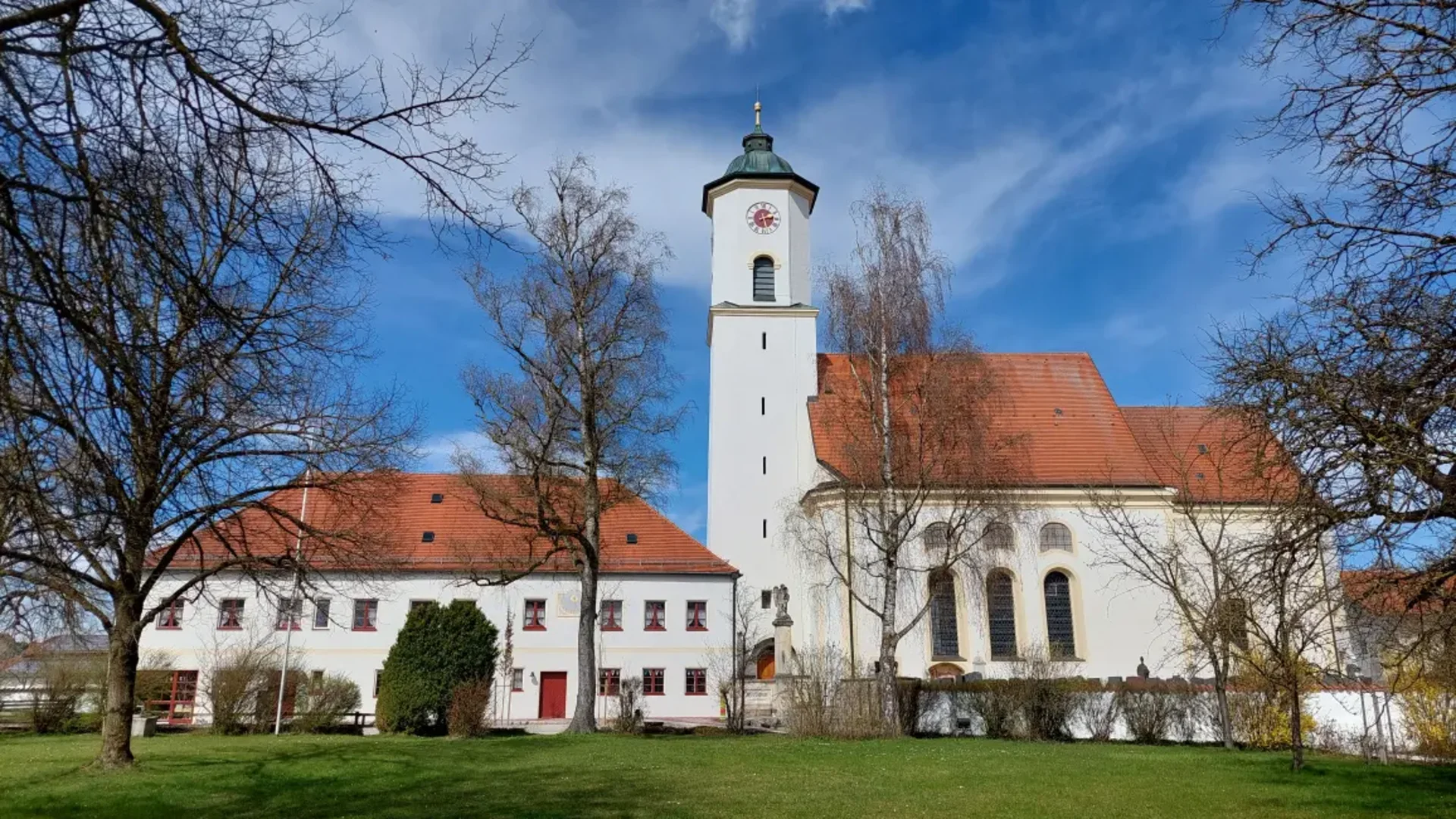 Weißer Kirchturm mit Uhr und rotem Ziegeldach im Grünen unter blauem Himmel
