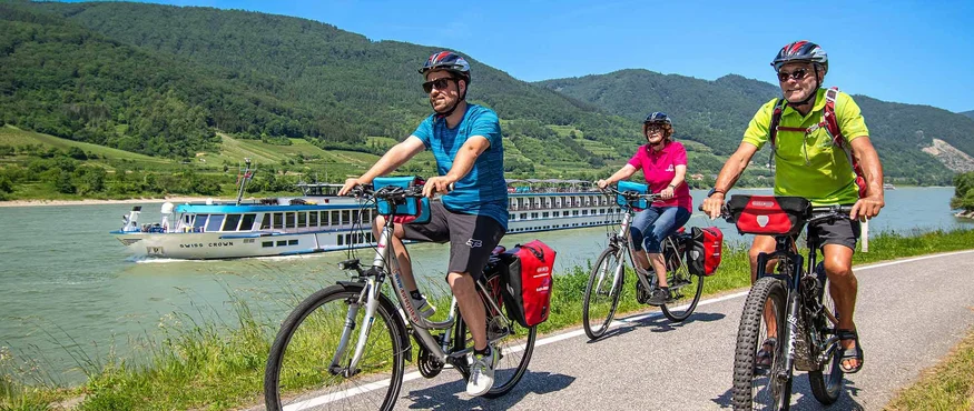 Three cyclists riding on a path beside a river with a cruise ship in the background