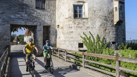 7 Quellen der Entschleunigung © Velontour - Heiko Mandl Zwei Radfahrer fahren über eine Holzbrücke an einer alten Burg vorbei bei klarem Himmel