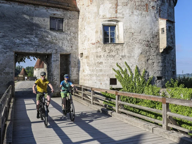 7 Quellen der Entschleunigung © Velontour - Heiko Mandl Zwei Radfahrer fahren über eine Holzbrücke an einer alten Burg vorbei bei klarem Himmel