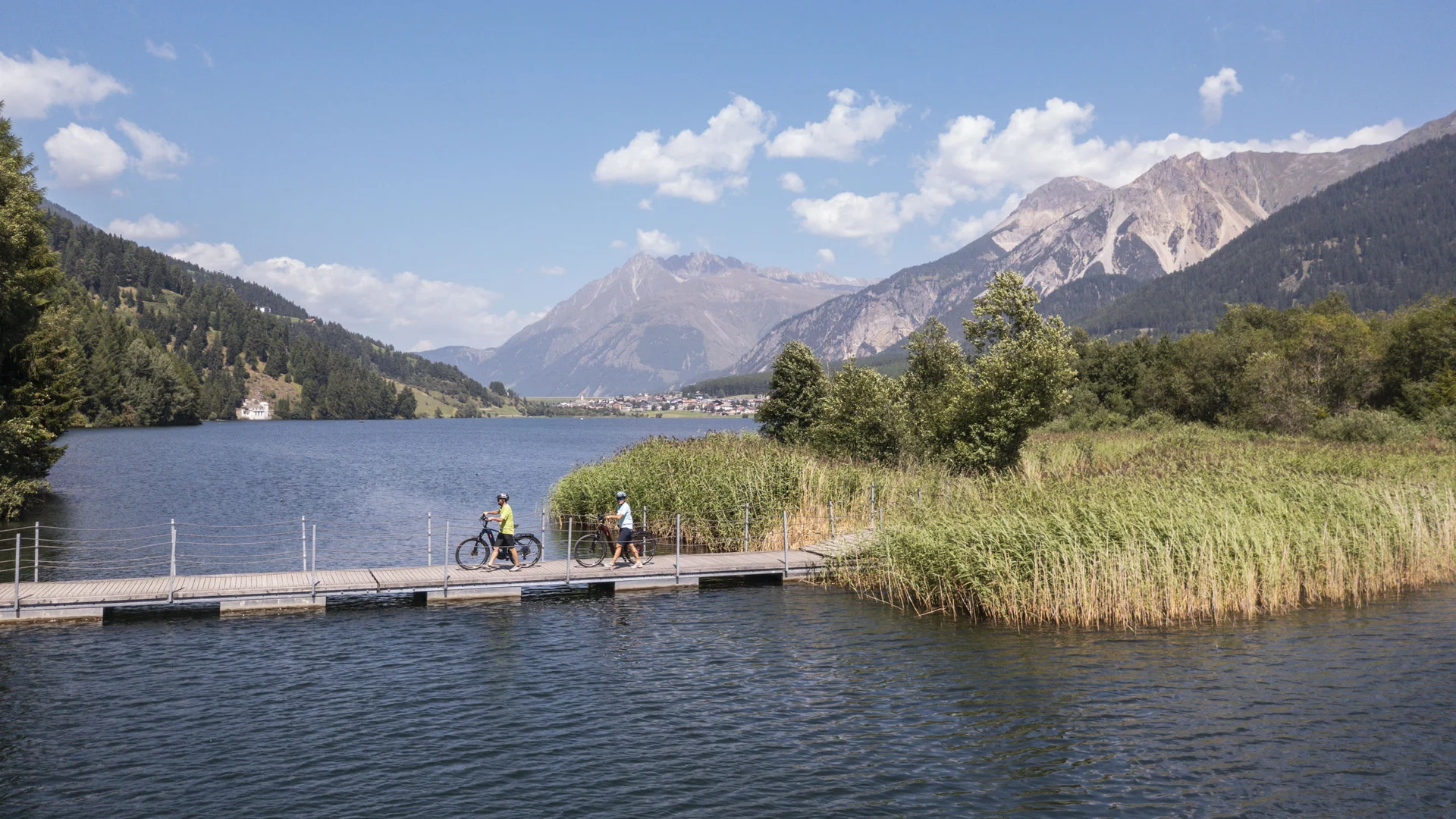 Zwei Radfahrer auf Steg an See mit Bergen und blauem Himmel im Hintergrund