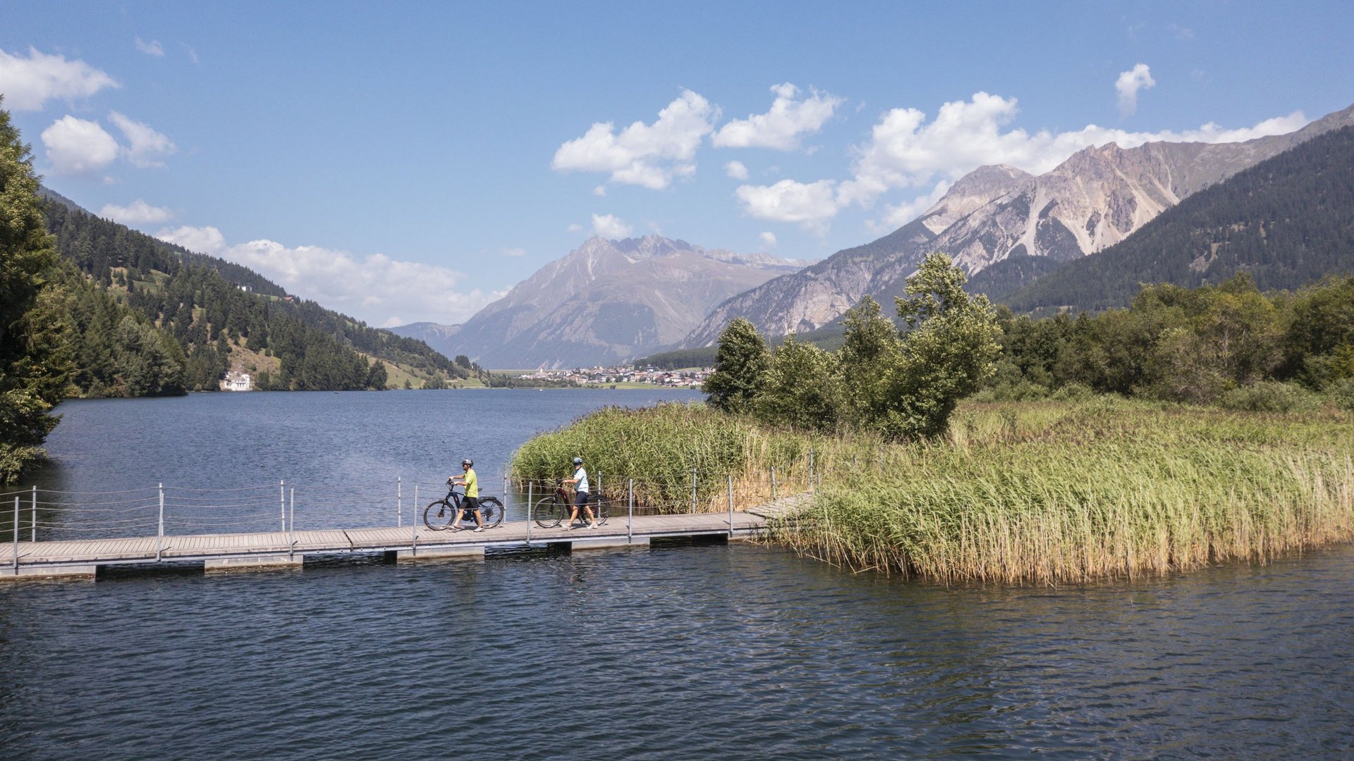 Two cyclists on a pier by a lake with mountains and blue sky in the background