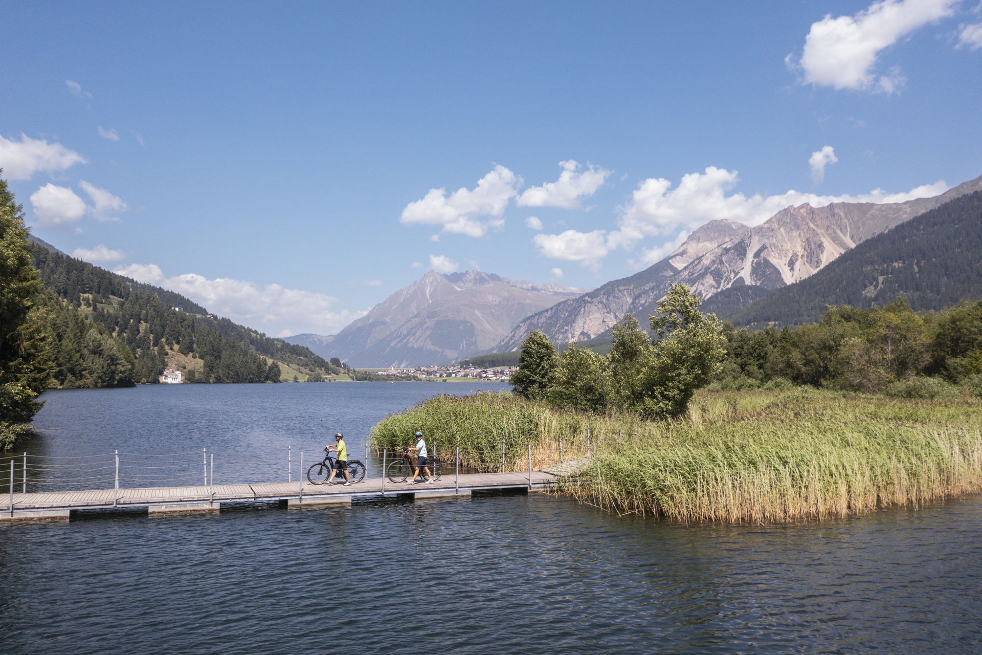 Two cyclists on a pier by a lake with mountains and blue sky in the background