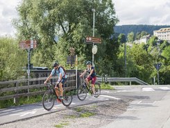 Two cyclists on a bike path in rural area with signs and mountains in background