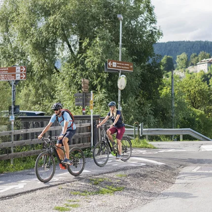 Twee fietsers op fietspad in landelijke omgeving met borden en bergen op achtergrond