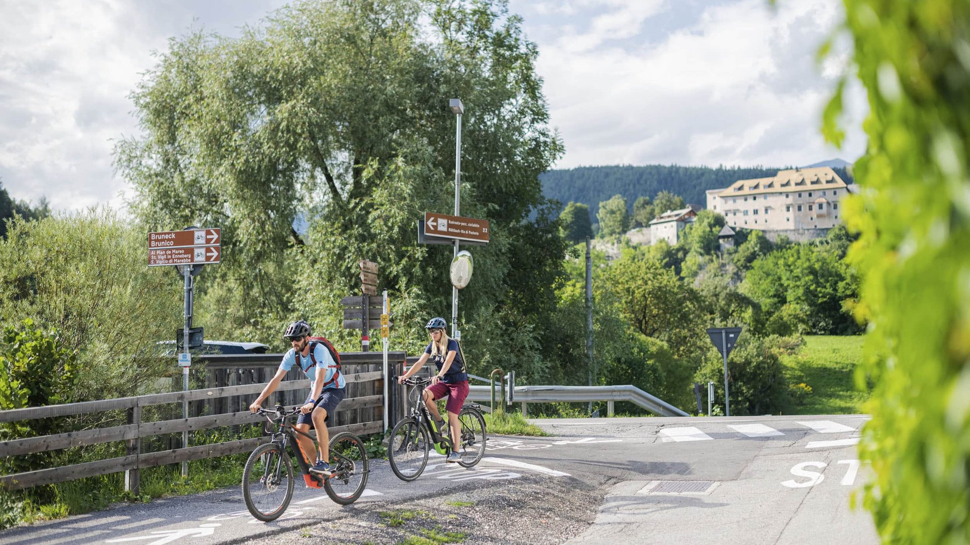 Two cyclists on a bike path in rural area with signs and mountains in background