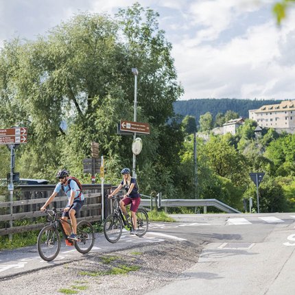 Two cyclists on a bike path in rural area with signs and mountains in background