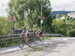 Twee fietsers op fietspad in landelijke omgeving met borden en bergen op achtergrond