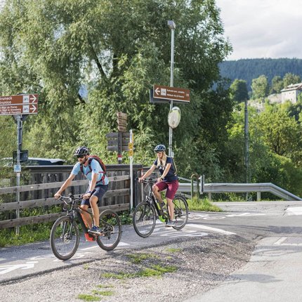 Two cyclists on a bike path in rural area with signs and mountains in background
