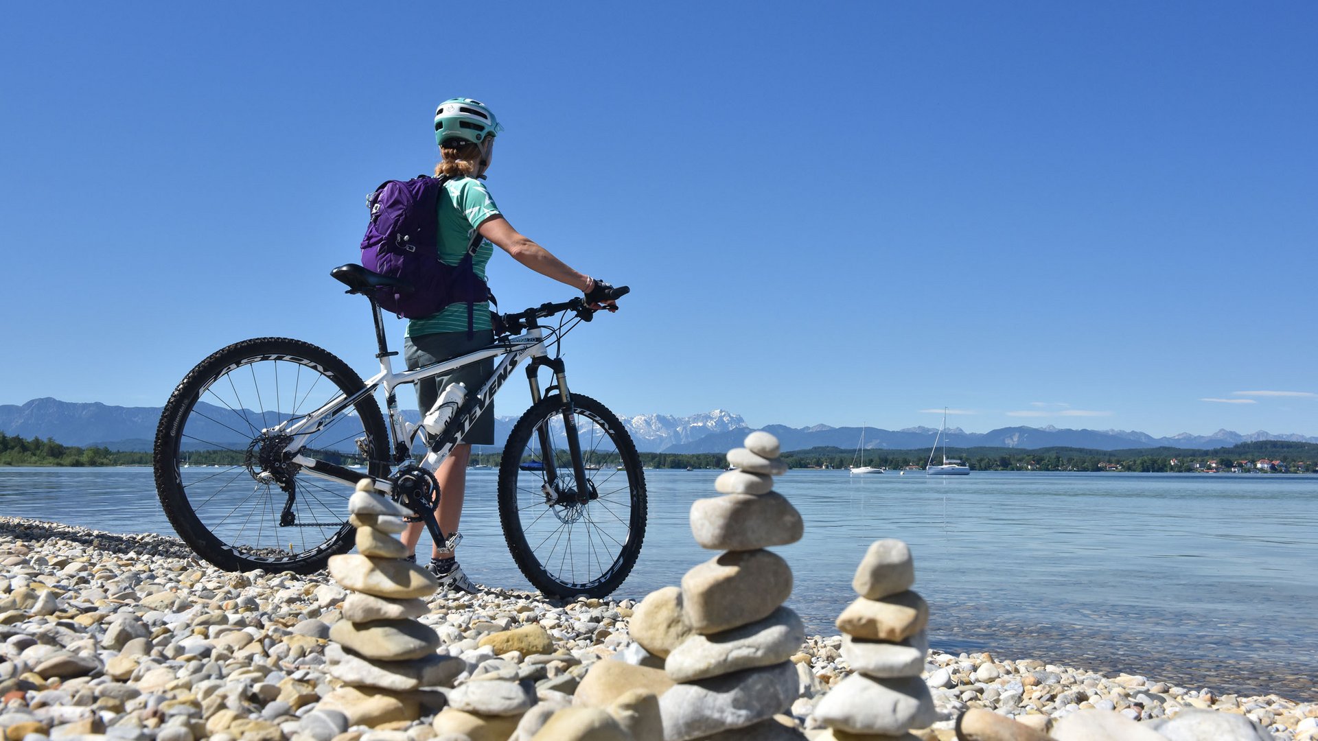 Woman with bike looking at stone stacks by lake with mountains in background