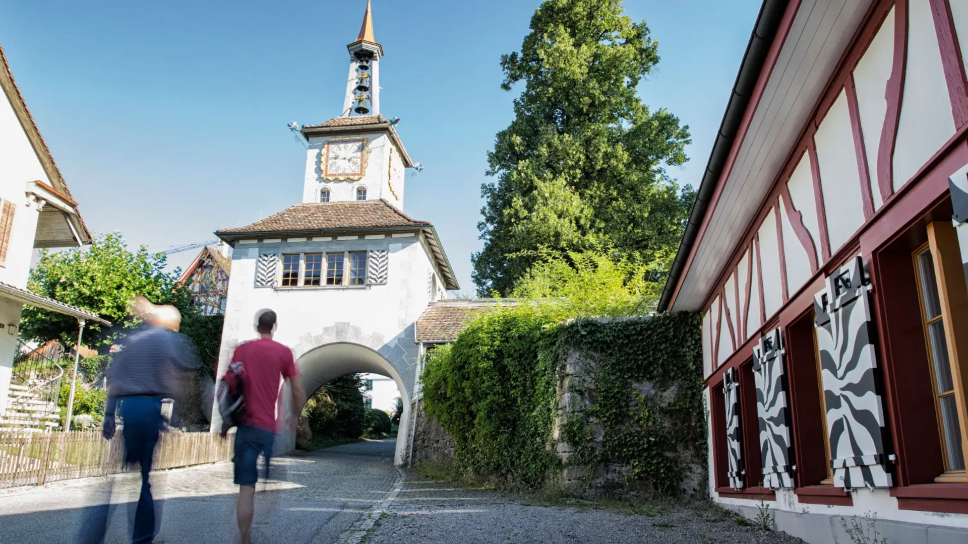 Two people walking towards a historic tower gate in a village