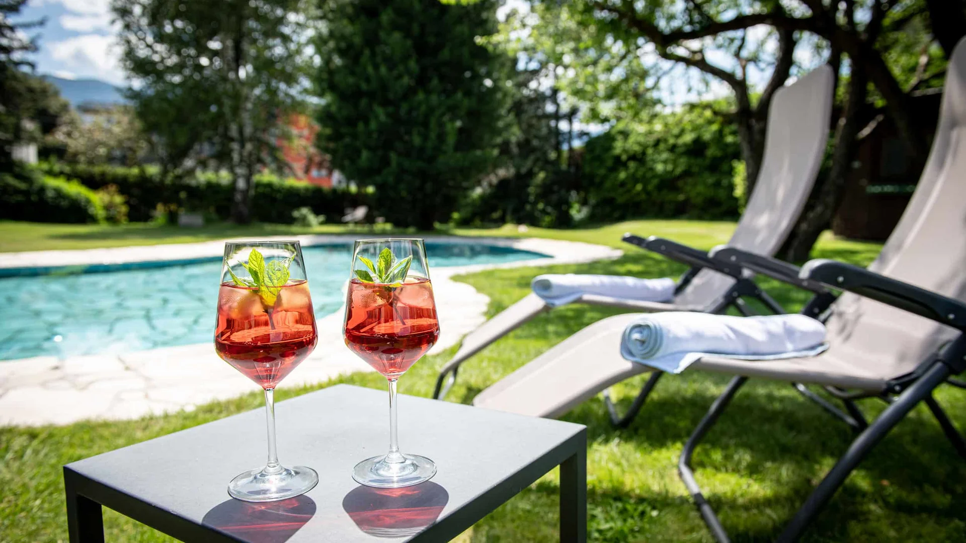 Two drinks on table near pool with lounge chairs in garden