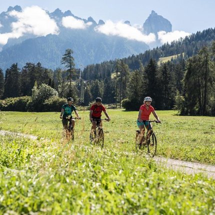 Three cyclists riding on a path through green meadow with mountains and clouds behind