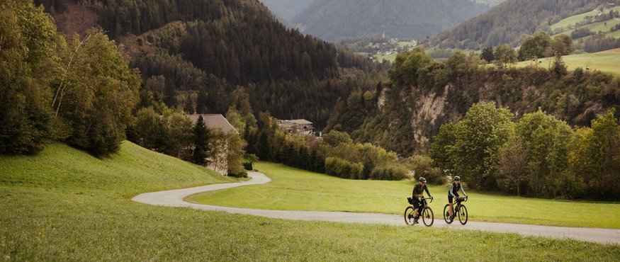 Two cyclists on rural road in green mountain landscape under cloudy sky