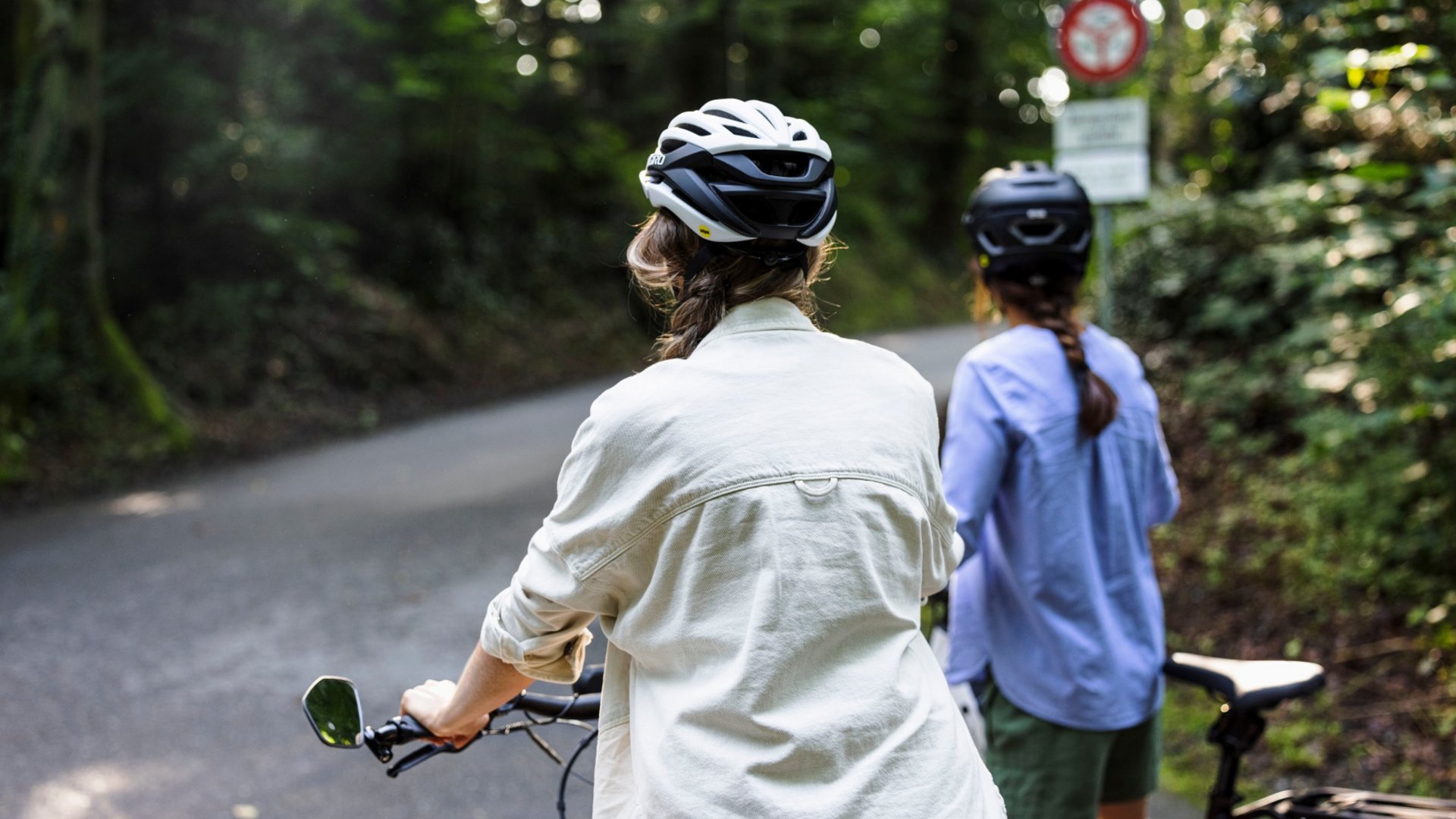 Two women wearing helmets stand with bicycles on a forest path