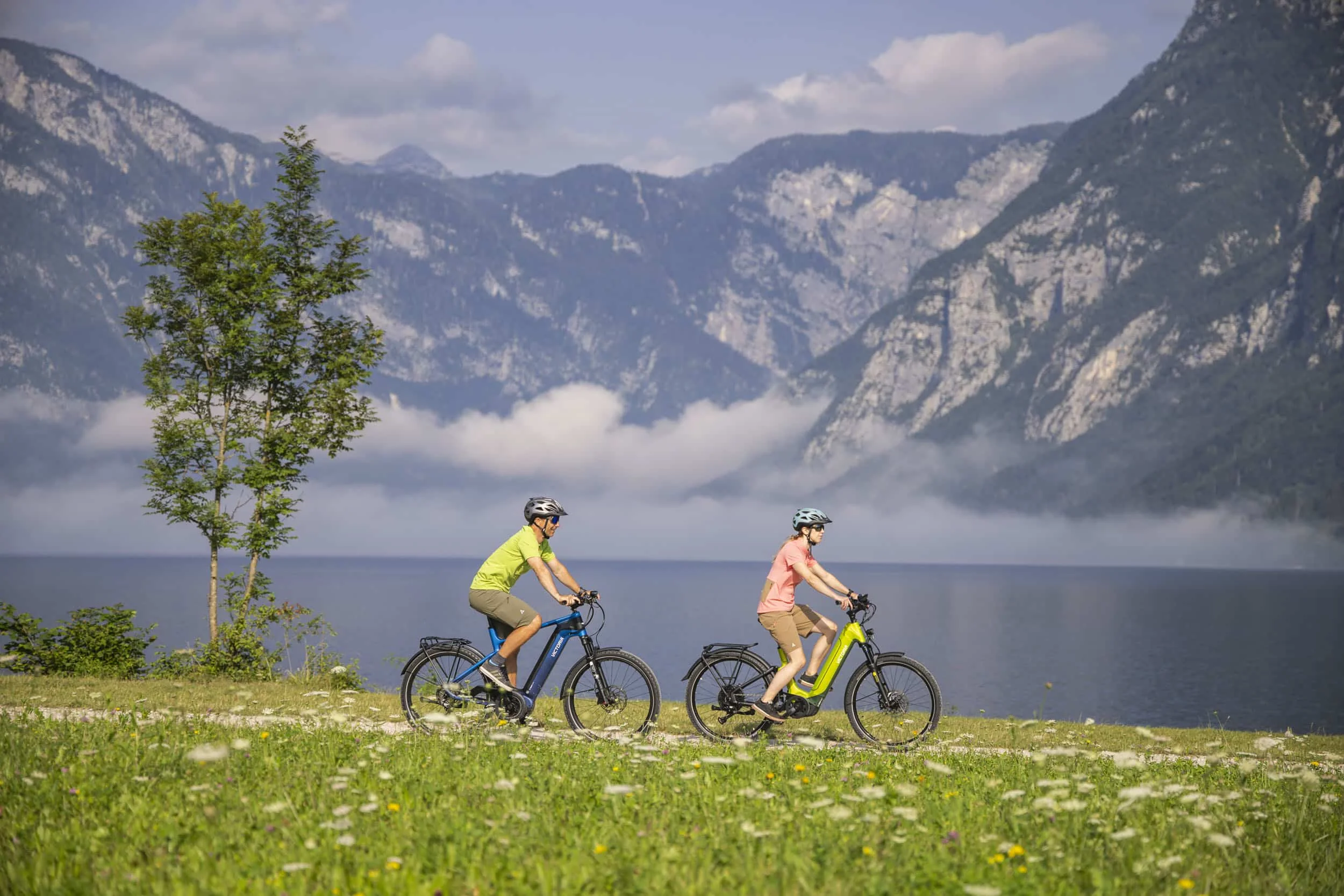 Zwei Radfahrer mit Helmen fahren an einem See mit Bergen im Hintergrund
