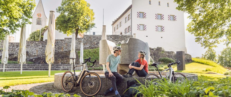 Zwei Radfahrer mit Helmen sitzen an Brunnen vor Schloss und Kirche