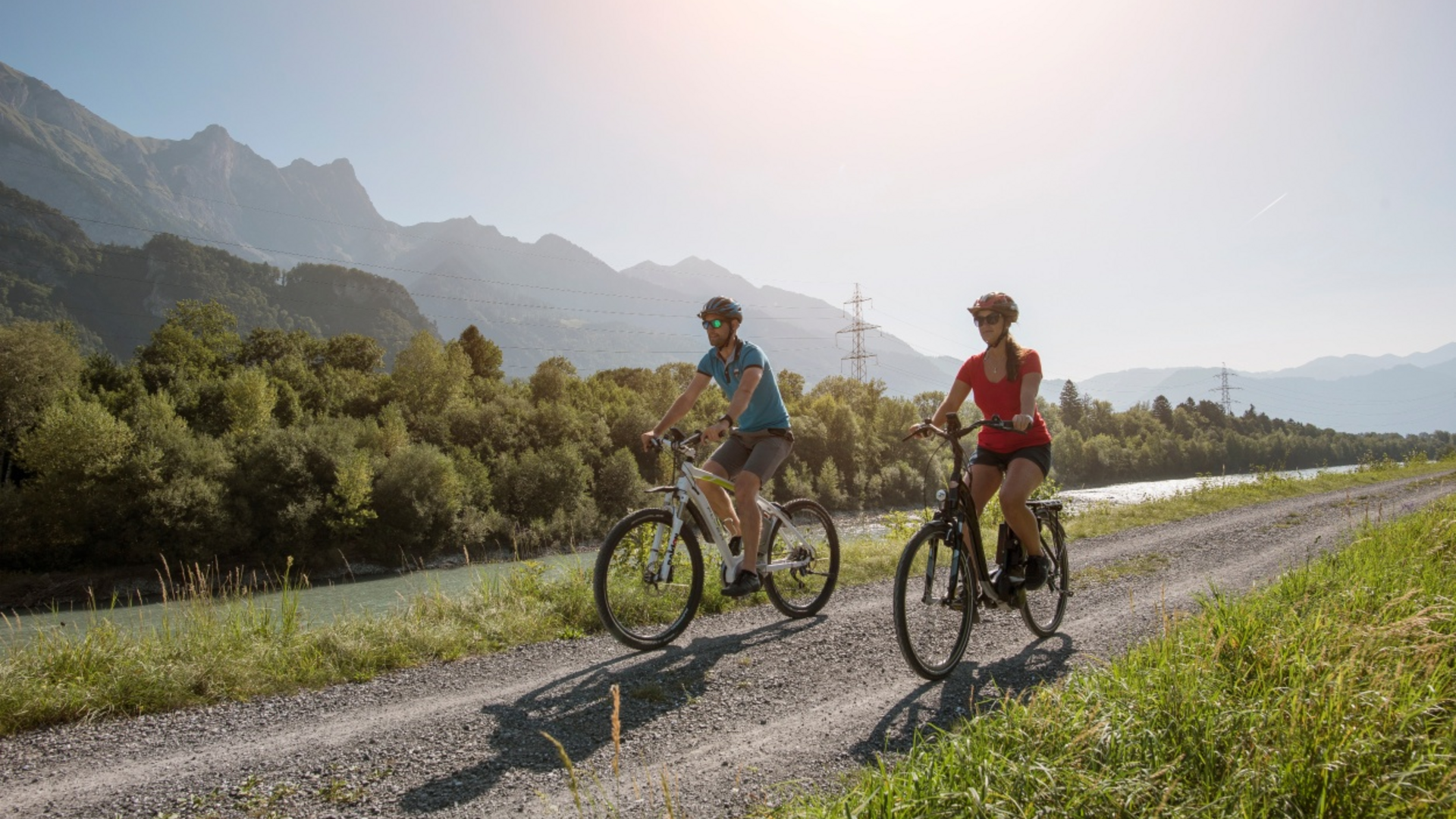Two people cycling on a path beside a river in the mountains