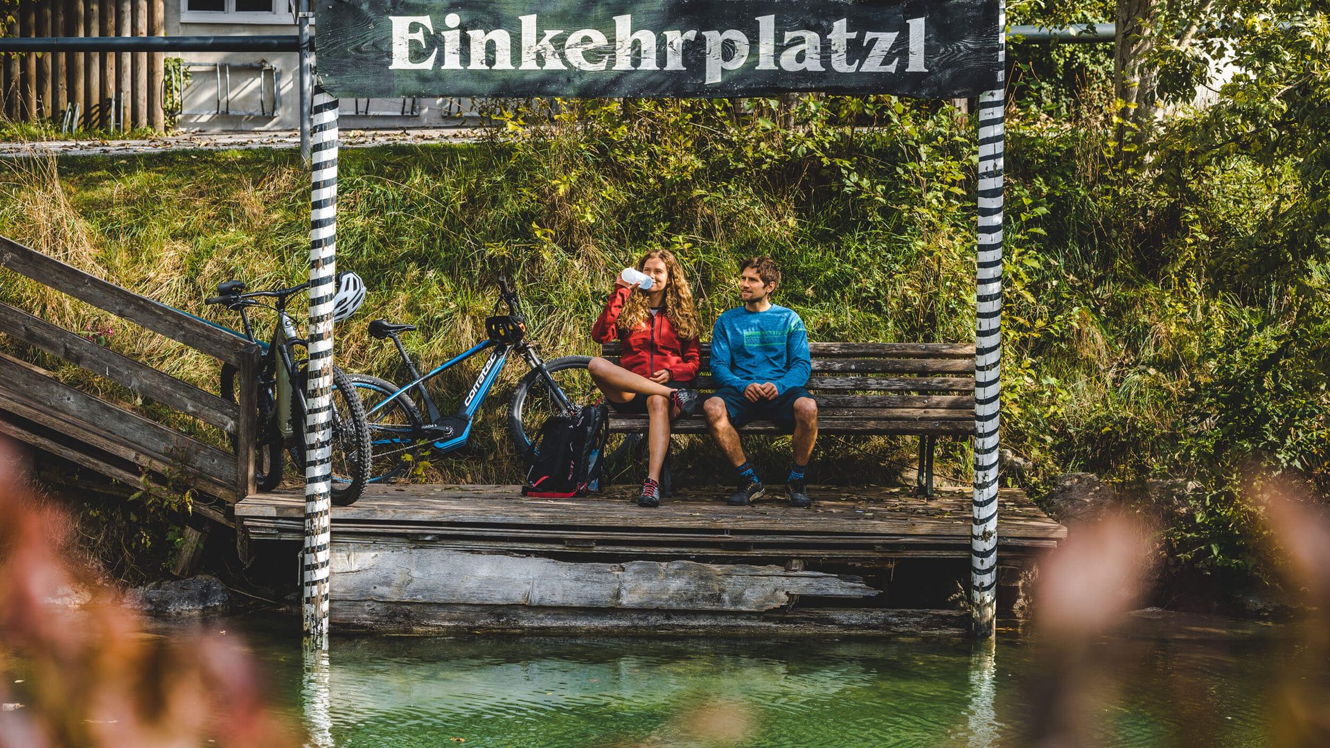 Two cyclists resting on a bench by the lake under a sign reading Einkehrplatzl