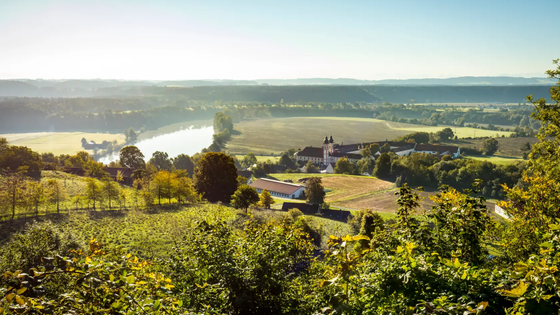Landscape with river, fields, and buildings under sunlight
