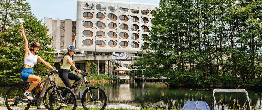Two women cycling by a lake near a modern building