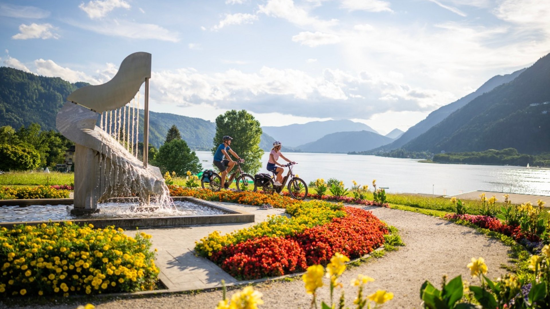 Cyclists ride along a flower-lined lake path with mountains in the background