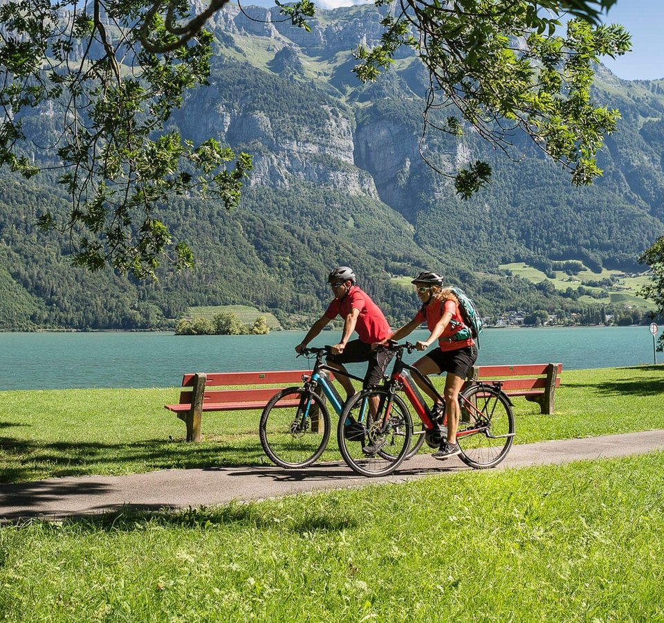 Zwei Radfahrer fahren am See vor Bergen auf einem sonnigen Radweg