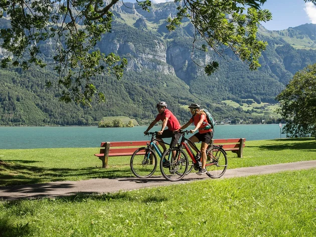 Zwei Radfahrer fahren am See vor Bergen auf einem sonnigen Radweg