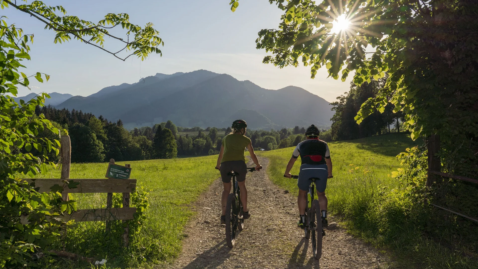 Im Sattel durchs Tölzer Land entlang von Isar & Loisach © Tölzer Land Tourismus - Bernd Ritschel Zwei Radfahrer auf einem Waldweg bei Sonnenschein, umgeben von Bäumen und Bergen