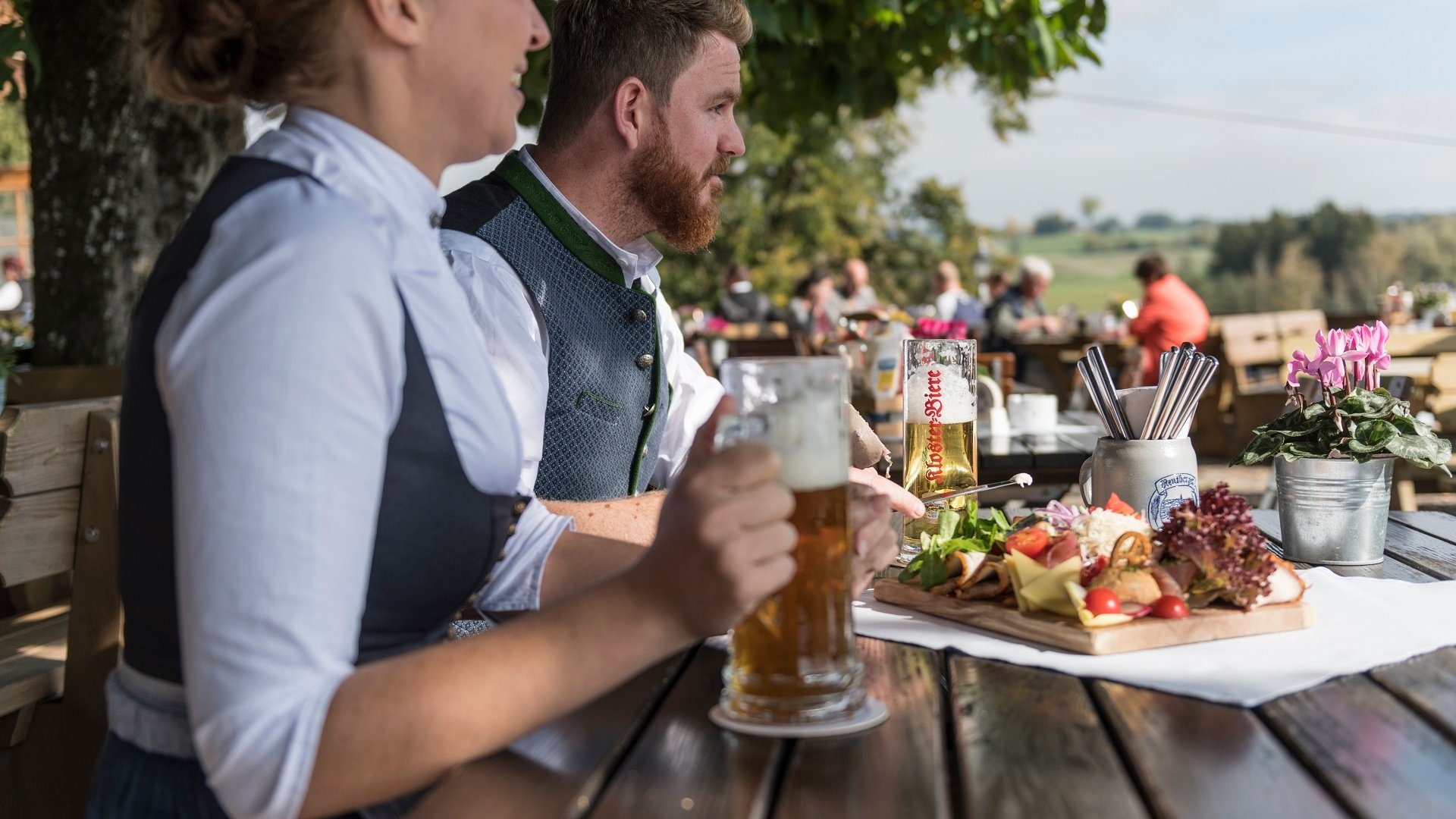 Two people in traditional clothing drinking beer at beer garden with snacks