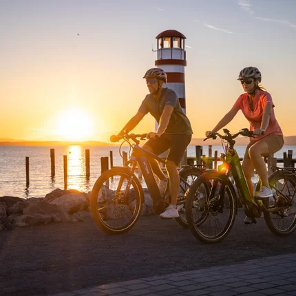 Zwei Radfahrer fahren bei Sonnenuntergang am Leuchtturm am Wasser