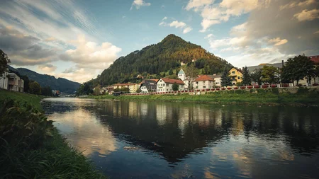 Flussufer mit Dorf und bewaldetem Berg im Hintergrund bei bewölktem Himmel