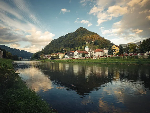 Flussufer mit Dorf und bewaldetem Berg im Hintergrund bei bewölktem Himmel