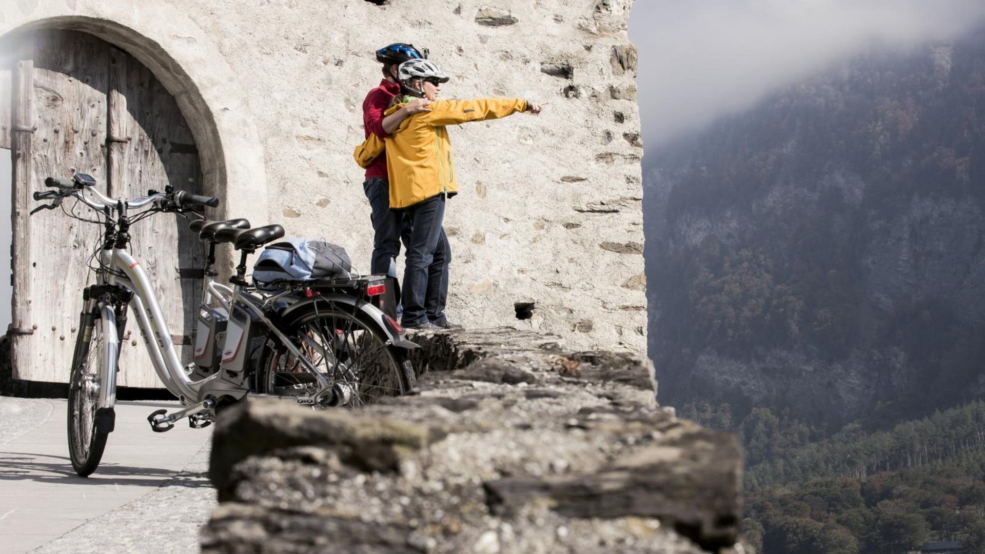 Two cyclists in helmets looking over castle wall at foggy valley