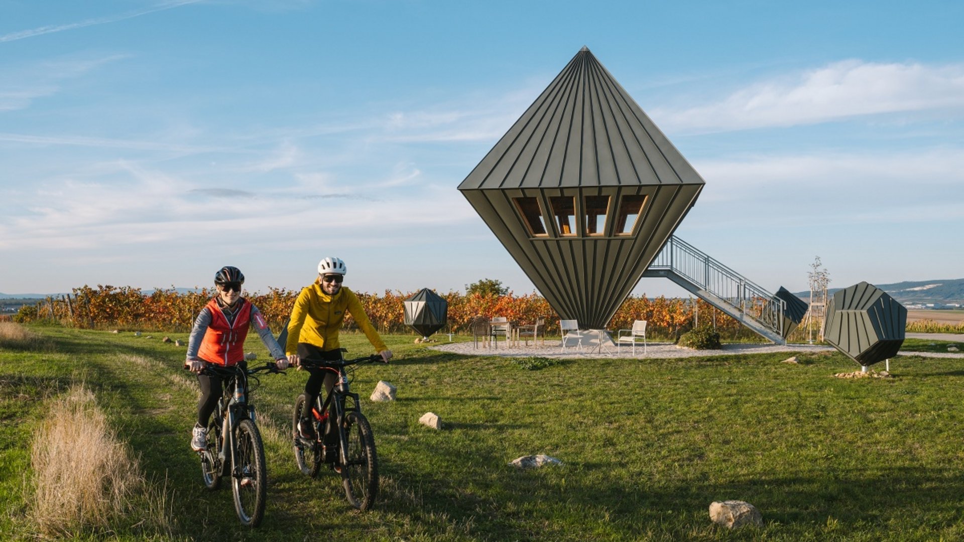 Two cyclists on a path near modern pointed treehouses outdoors